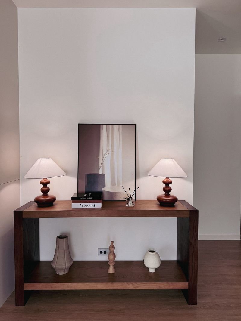Horizon_By_Patta_13-5mb_Dlh538_7 Wood console table with two matching white-shaded lamps flanking a framed image, against a white wall.