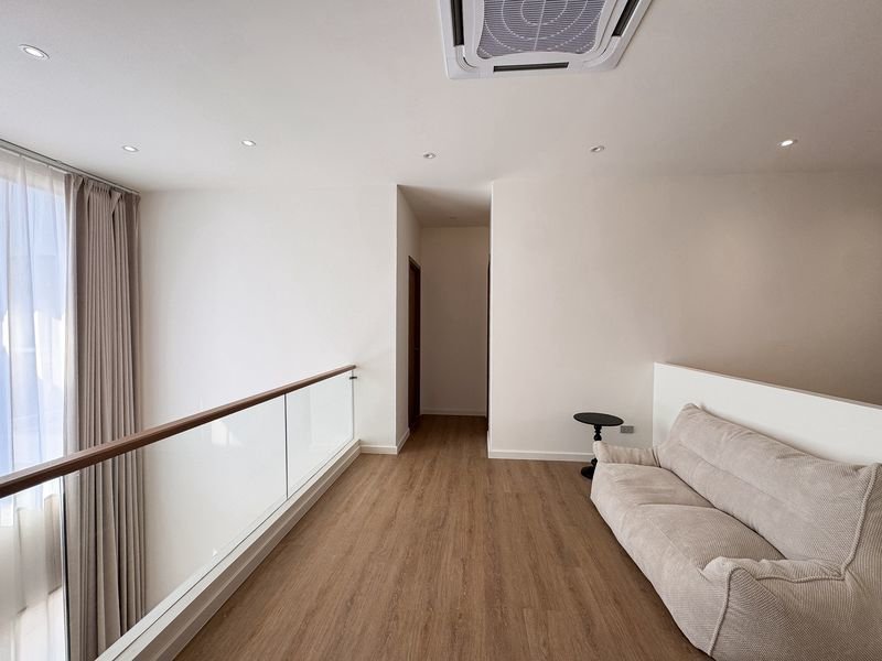 Bright loft hallway with a beige sofa on the right, glass railing on the left, and a doorway at the far end.