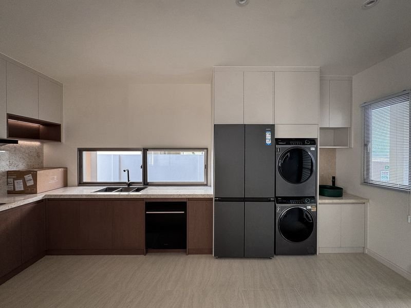Modern kitchen with a stainless steel fridge and a stacked front-load washer and dryer beside white upper cabinets and light wood lower cabinets.