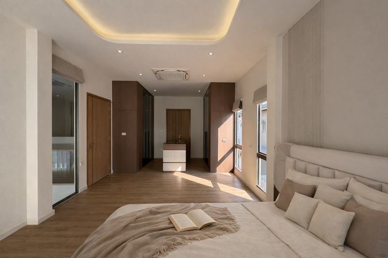 Modern bedroom with a large headboard bed, beige pillows, and warm natural light streaming through windows.