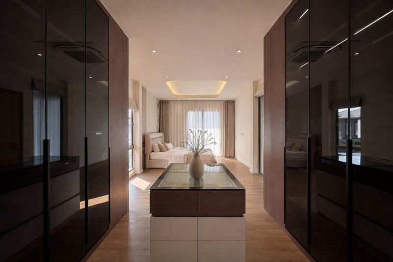 Luxurious hotel-style master suite with dark glossy wardrobes lining a central island and a vase on top, at the end of a hallway leading to a bed area.