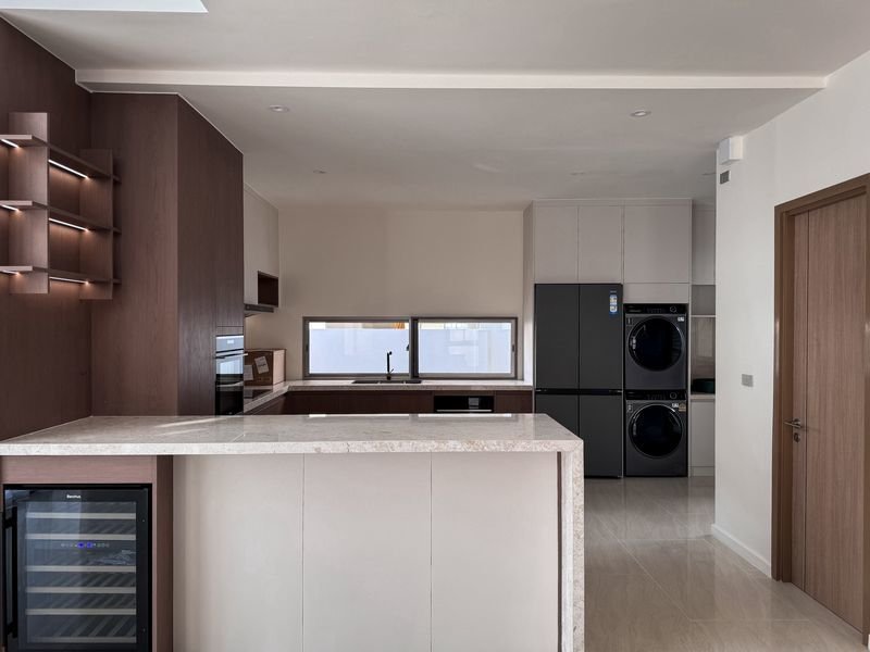 Modern kitchen with a beige island, dark wood cabinets, a windowed sink area, and a stainless steel fridge; stacked washer and dryer in the background.