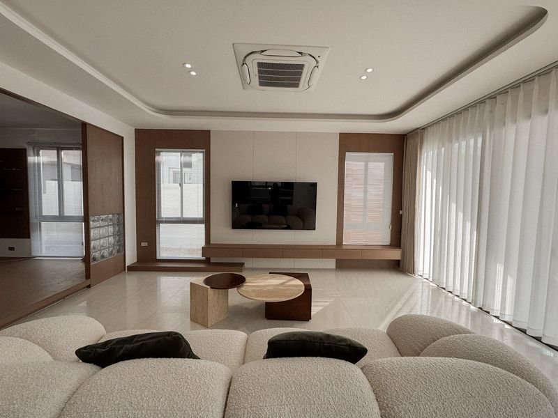 Bright modern living room with a wall-mounted TV, low wooden media bench, and a white curved sofa facing the screen; floor-to-ceiling curtains on the right.