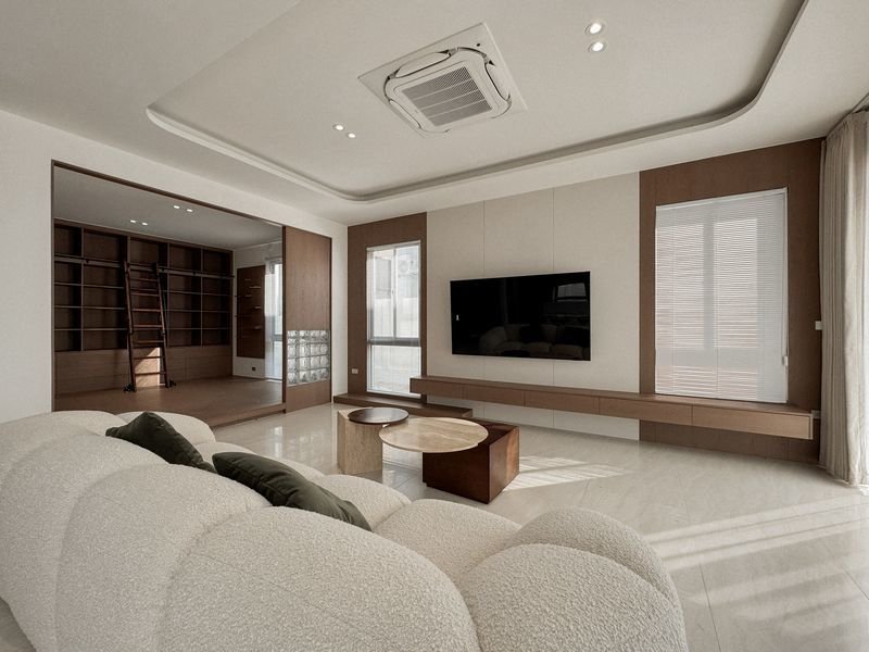 Modern living room featuring a curved white sofa facing a wall-mounted TV, glass coffee tables, and built-in wood shelves; bright with large windows.