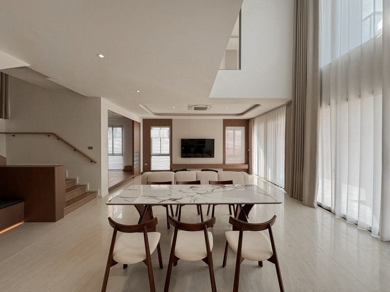 Modern open-concept dining area with a marble table and six wooden chairs, large windows with sheer curtains.