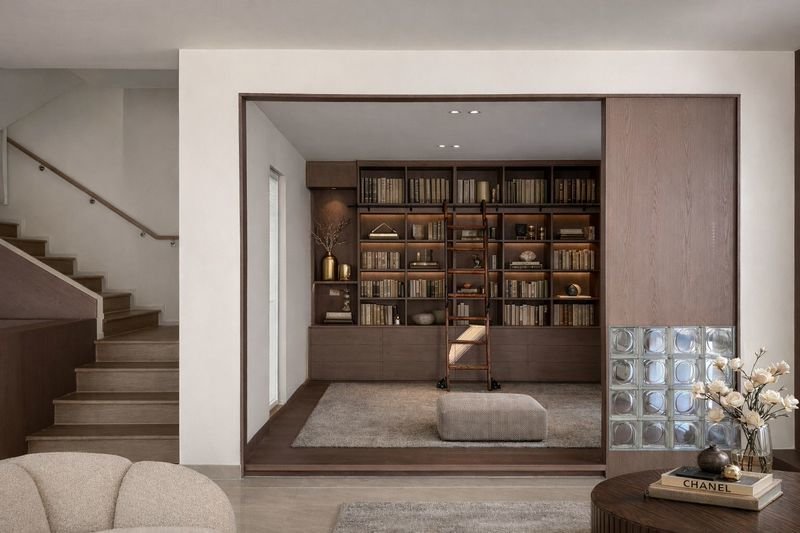 Hidden home library revealed behind a sliding wood wall, with floor-to-ceiling bookshelves, ladder, and cozy seating area in a modern living room.