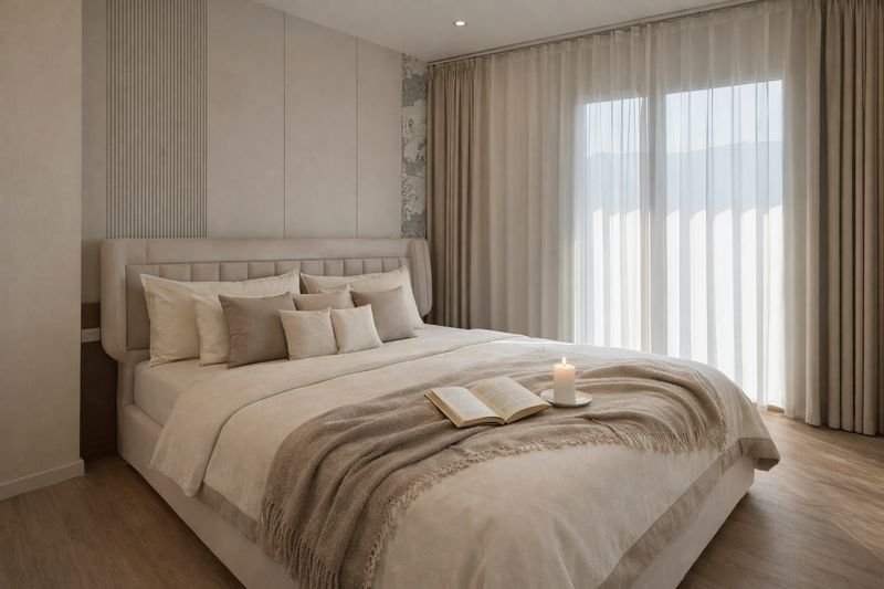Neutral luxury bedroom with a large upholstered bed, beige pillows, and an open book with a lit candle on a beige throw at the foot of the bed.