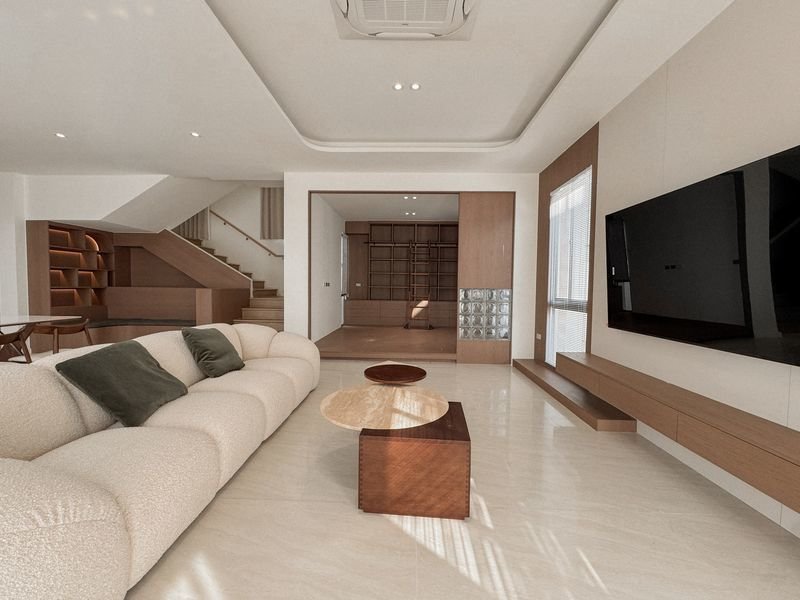 Modern living room with a beige sectional sofa, wooden coffee tables, and a wall-mounted flat-screen TV.