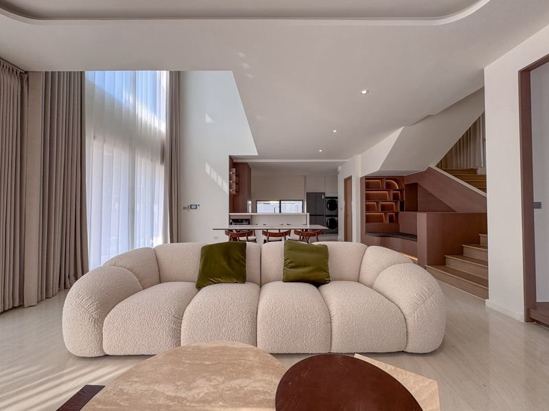 Open-plan living room with a cream curved sofa, two green cushions, tall windows with curtains, and a wooden staircase with built-in shelves in the background.