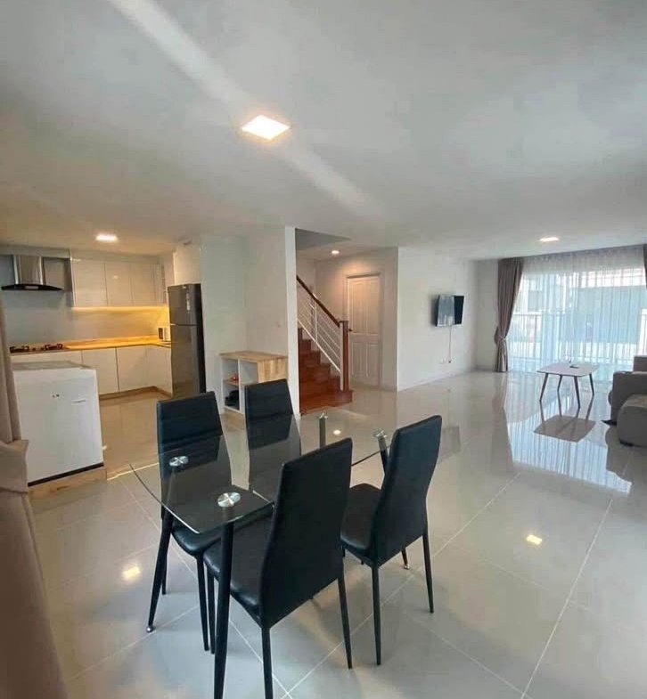 Open-plan living and dining area with a glass dining table and six black chairs, a modern kitchen to the left, and a staircase in the center.