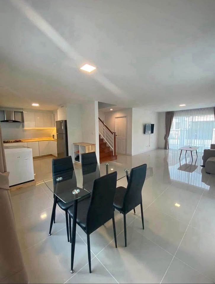 Open-plan living and dining area with a glass dining table and six black chairs, a modern kitchen to the left, and a staircase in the center.