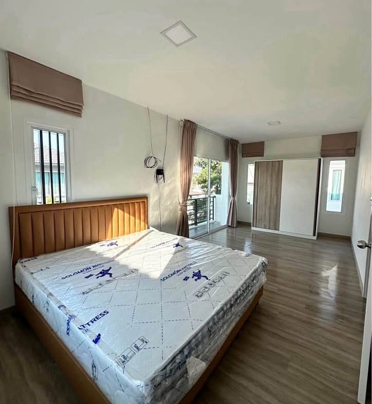 Bedroom with a wooden bed frame and a new mattress still wrapped in plastic, near a balcony door and wardrobe.