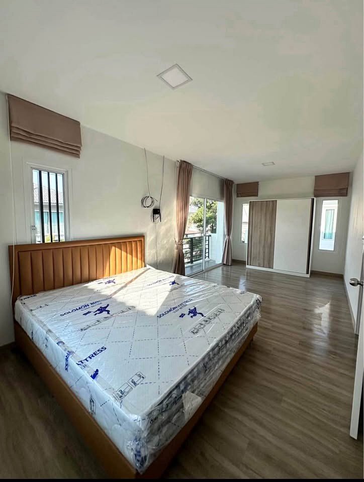 Bedroom with a wooden bed frame and a new mattress still wrapped in plastic, near a balcony door and wardrobe.