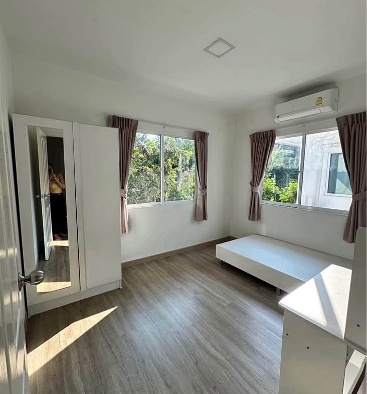 Bright furnished bedroom with a white bed, dresser, and wardrobe; beige curtains on two windows and a wall-mounted AC unit above.