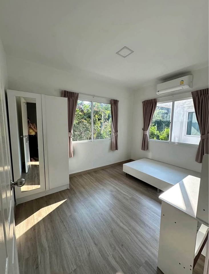 Bright furnished bedroom with a white bed, dresser, and wardrobe; beige curtains on two windows and a wall-mounted AC unit above.