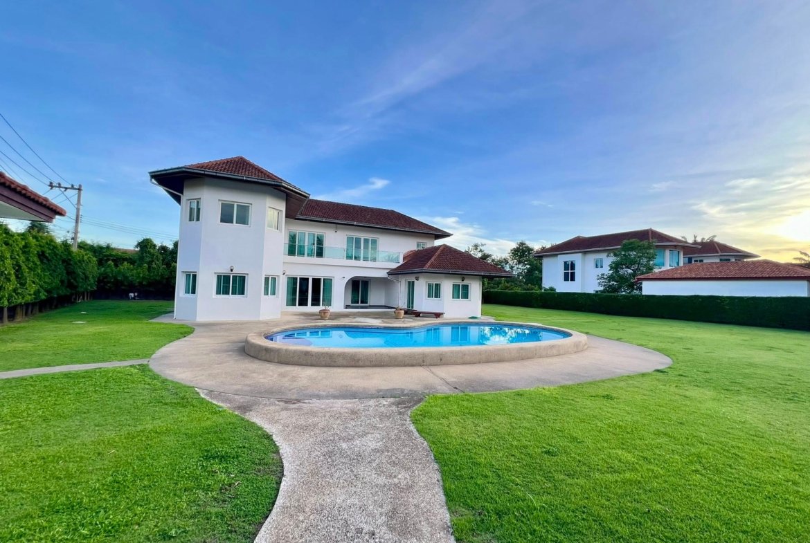Modern white, two-story house with a red-tiled roof, large windows, and a circular in-ground pool in the lawn beneath a clear blue sky.