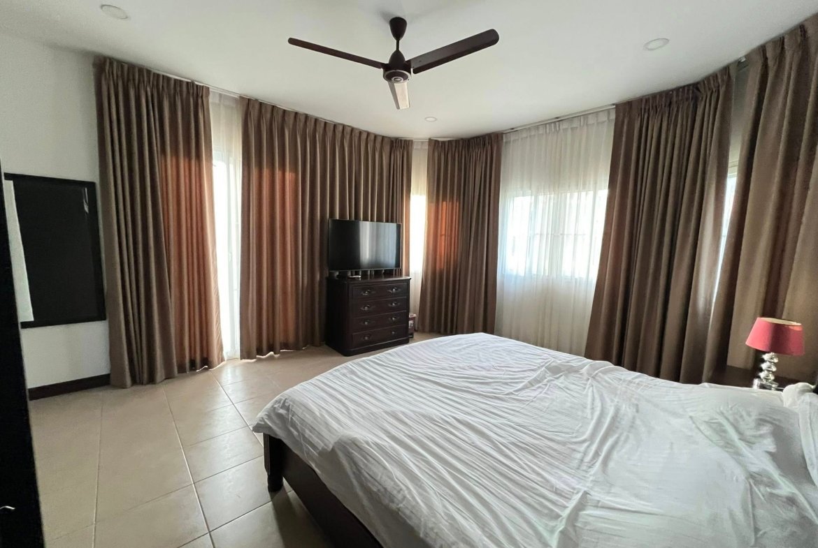 Bedroom with white bedspread in foreground, dark wood dresser with TV, and floor-to-ceiling brown curtains.