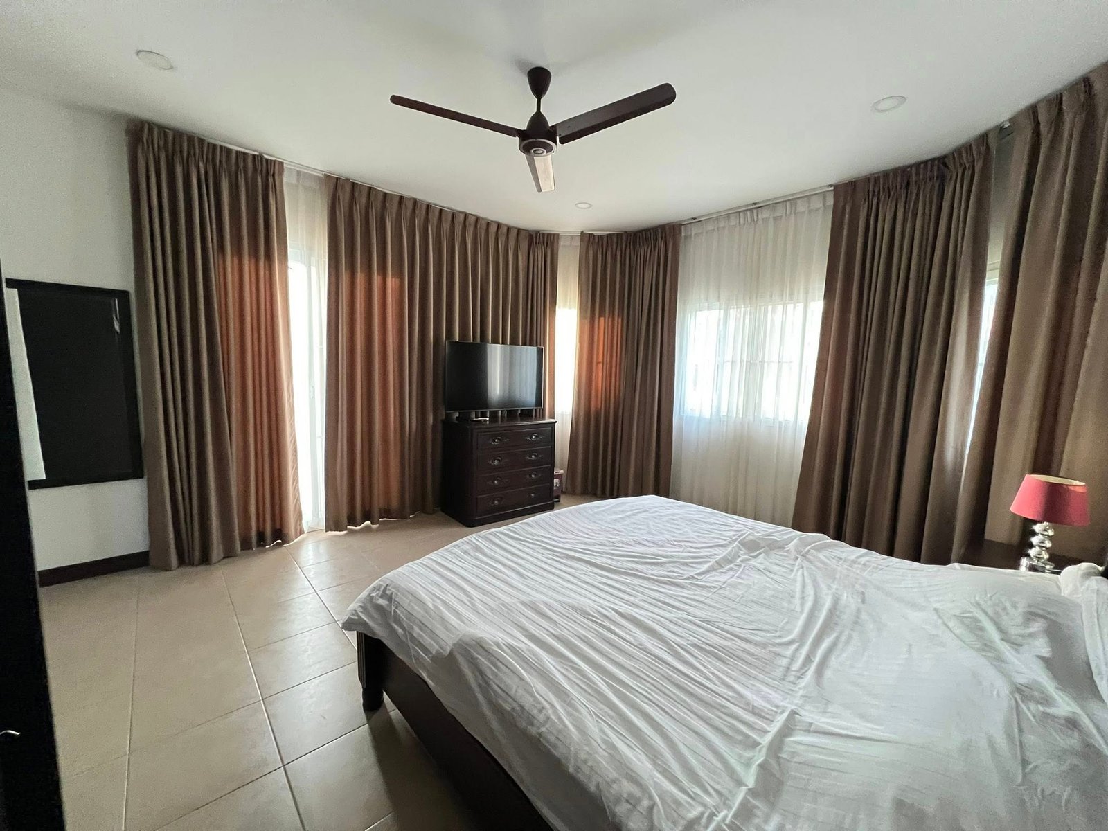 House_Sale_Mabprachan_Pattaya_Dlh533_11 Bedroom with white bedspread in foreground, dark wood dresser with TV, and floor-to-ceiling brown curtains.