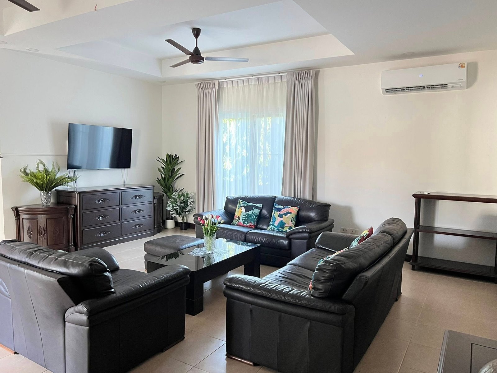 House_Sale_Mabprachan_Pattaya_Dlh533_14 Living room with black leather sectional, wall-mounted TV on a dark dresser, and potted plants.