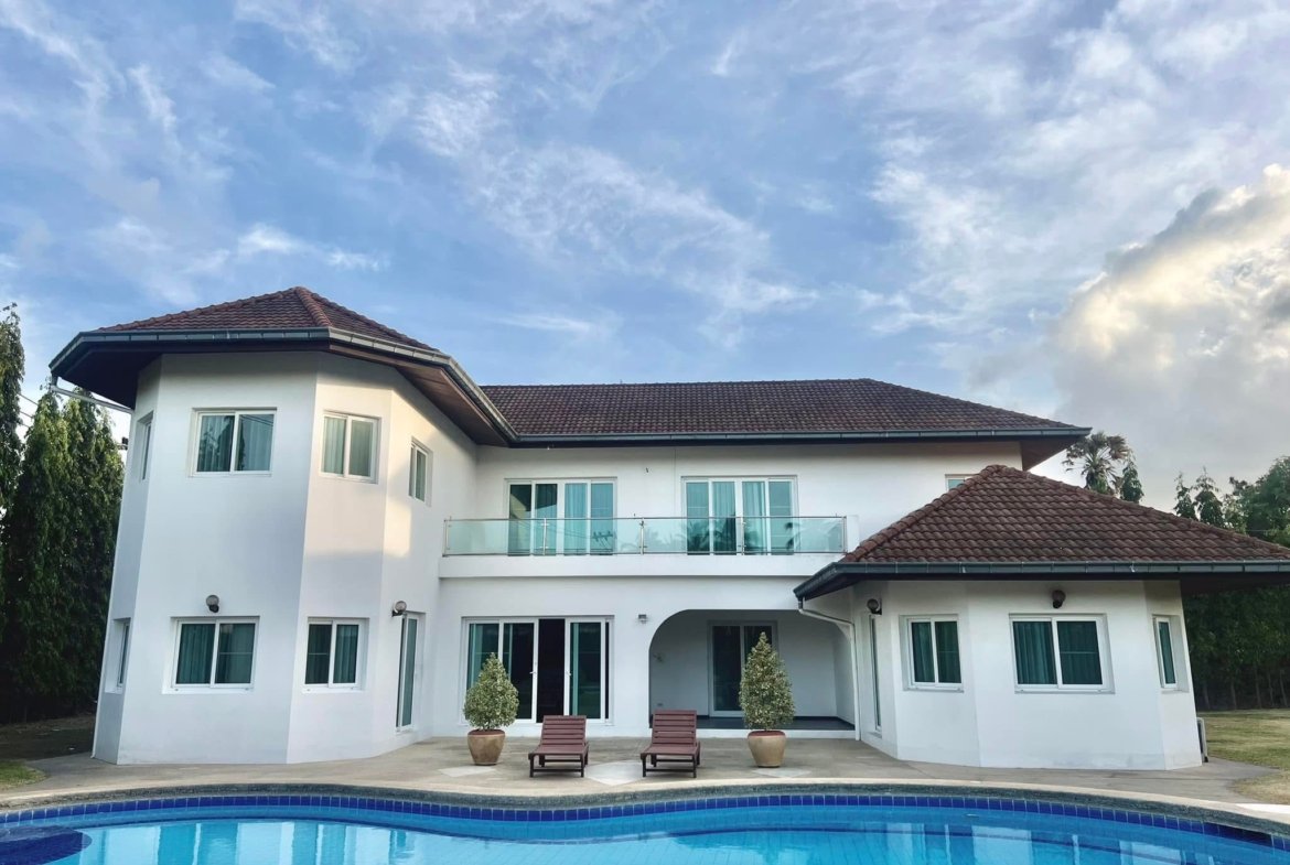Modern white house with red-tiled roof, glass balcony, and a blue swimming pool in the foreground.
