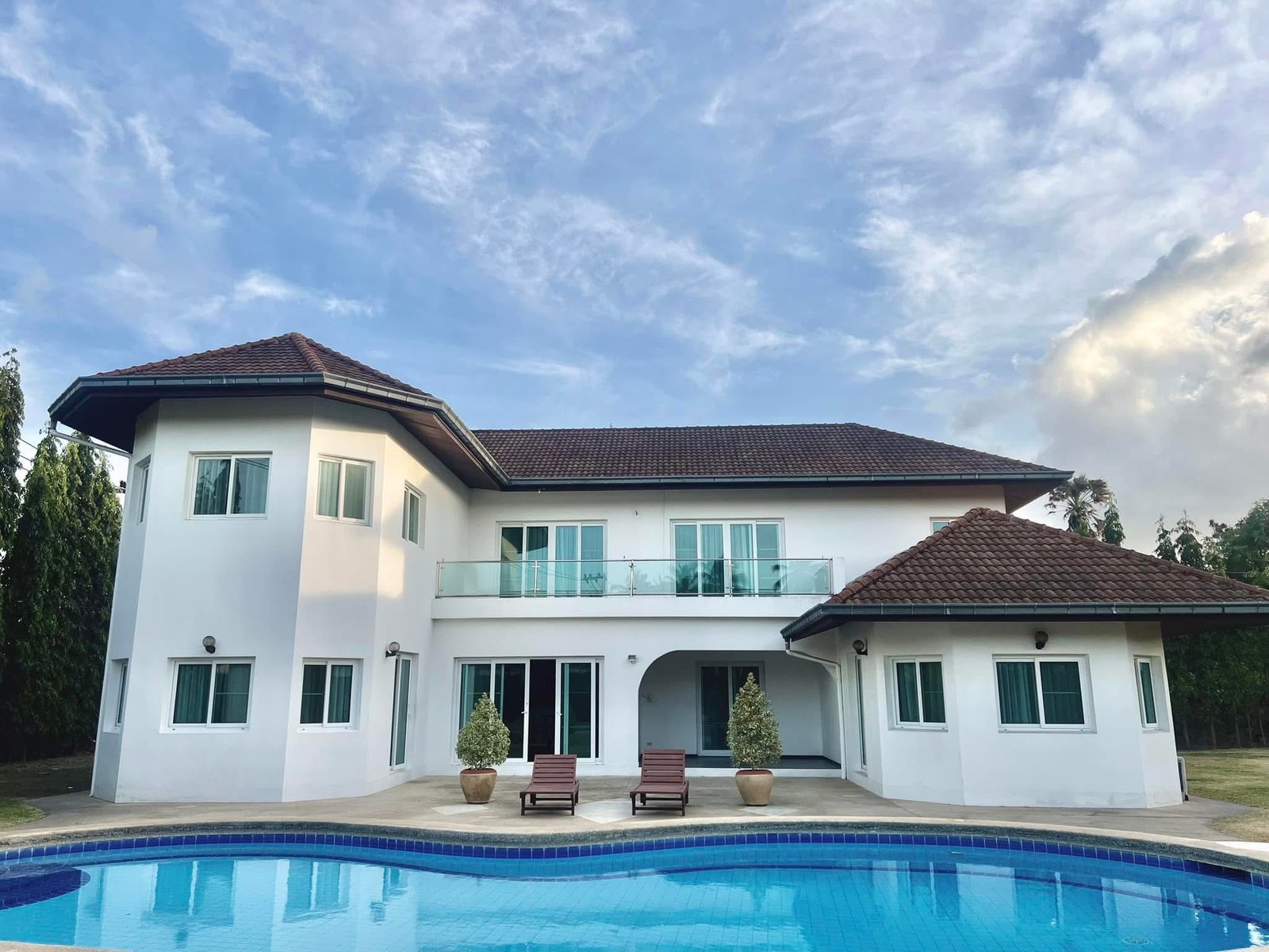 House_Sale_Mabprachan_Pattaya_Dlh533_2 Modern white house with red-tiled roof, glass balcony, and a blue swimming pool in the foreground.