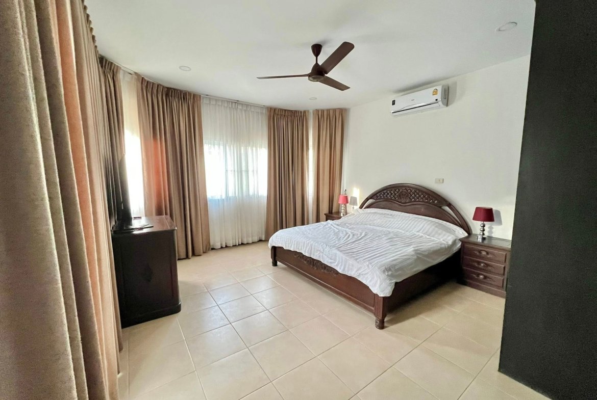 Hotel-style bedroom with a large wooden bed, matching nightstands, and beige curtains; ceiling fan overhead and wall-mounted A/C unit.
