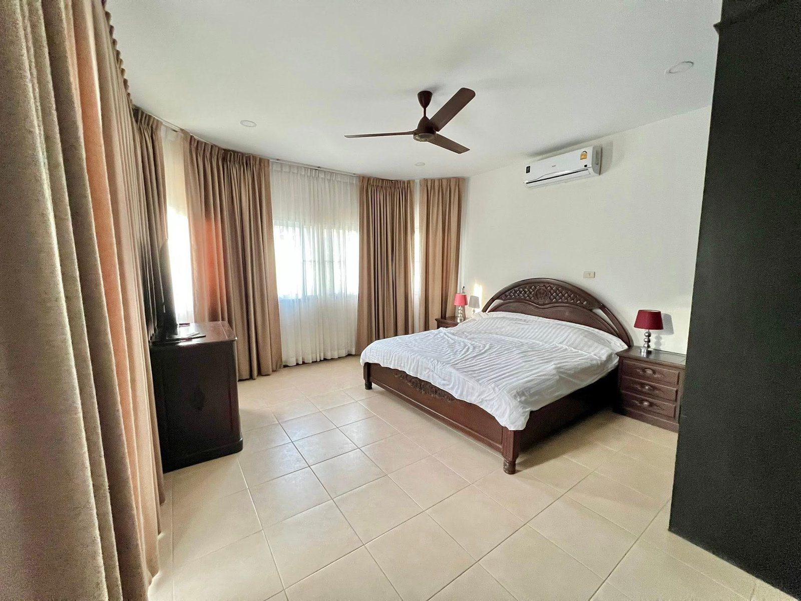 House_Sale_Mabprachan_Pattaya_Dlh533_20 Hotel-style bedroom with a large wooden bed, matching nightstands, and beige curtains; ceiling fan overhead and wall-mounted A/C unit.