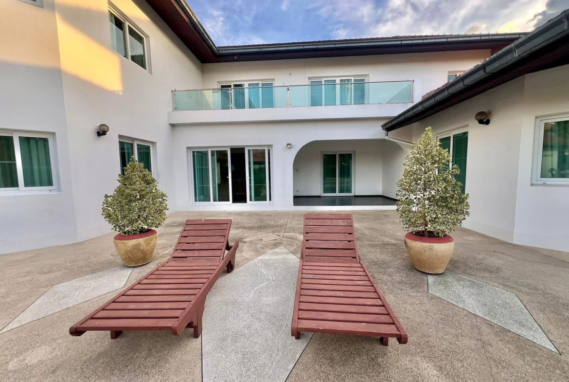 Courtyard of a modern white building with glass railing balcony, sliding doors, and two wooden lounge chairs nearby.