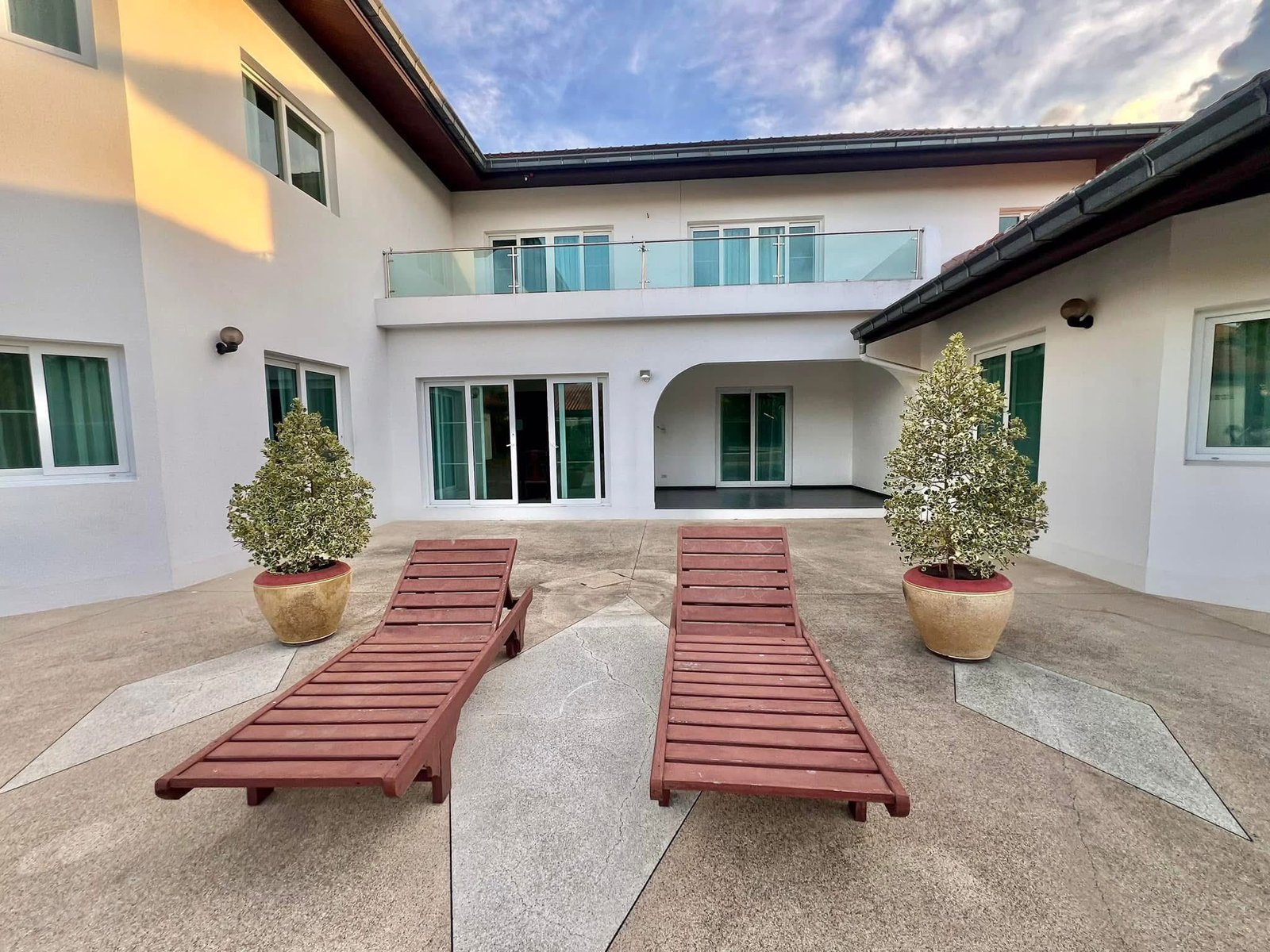 House_Sale_Mabprachan_Pattaya_Dlh533_23 Courtyard of a modern white building with glass railing balcony, sliding doors, and two wooden lounge chairs nearby.