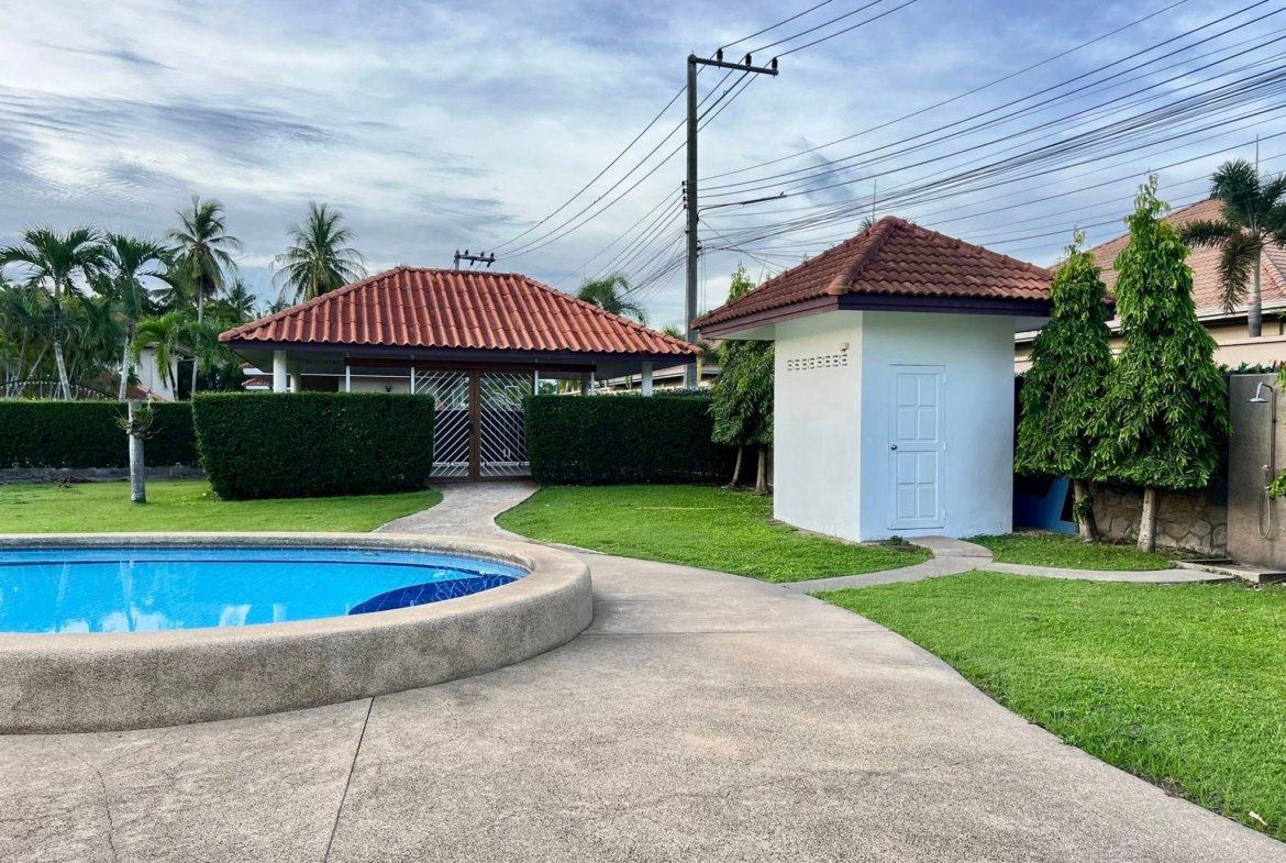 Pool edge and blue water curve in a backyard with a white building and red-tiled roof, palm trees in the background.