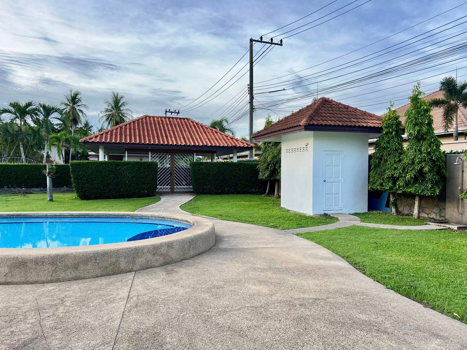 House_Sale_Mabprachan_Pattaya_Dlh533_24 Pool edge and blue water curve in a backyard with a white building and red-tiled roof, palm trees in the background.