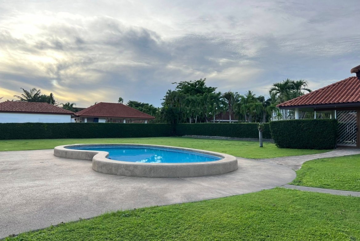 Circular blue pool set in a suburban yard with a curved concrete edge, green lawn, hedges, and red-tiled houses in the background under a cloudy sky.