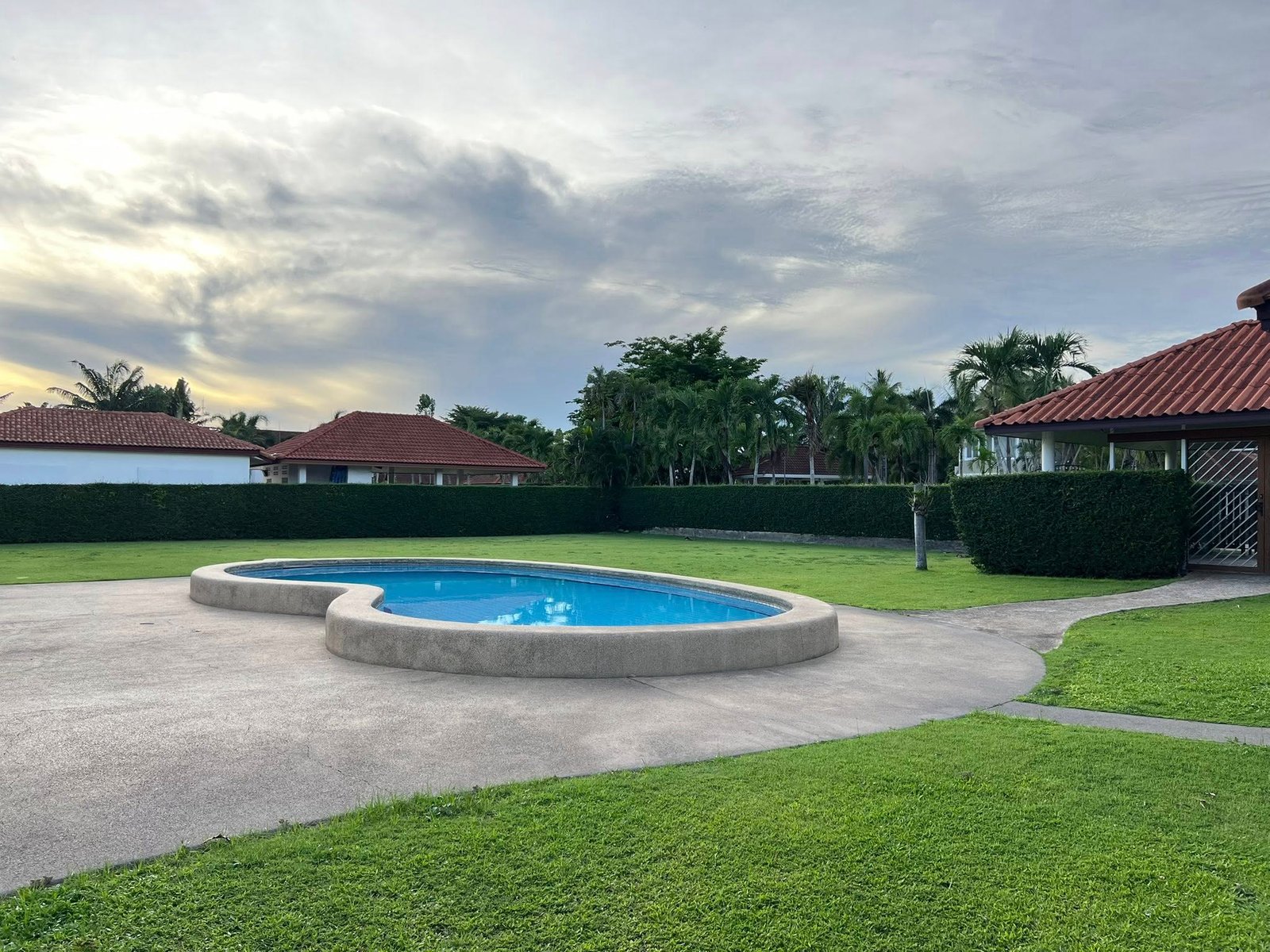 House_Sale_Mabprachan_Pattaya_Dlh533_25 Circular blue pool set in a suburban yard with a curved concrete edge, green lawn, hedges, and red-tiled houses in the background under a cloudy sky.