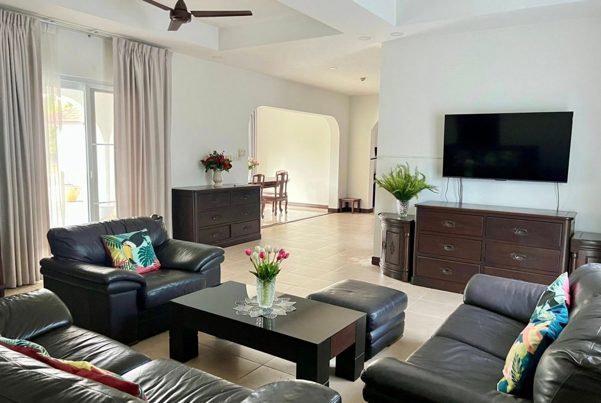 Bright living room with black leather sectional, glass-top coffee table, and multiple throw pillows; TV on wall to the right, plants and dark wood furniture throughout.
