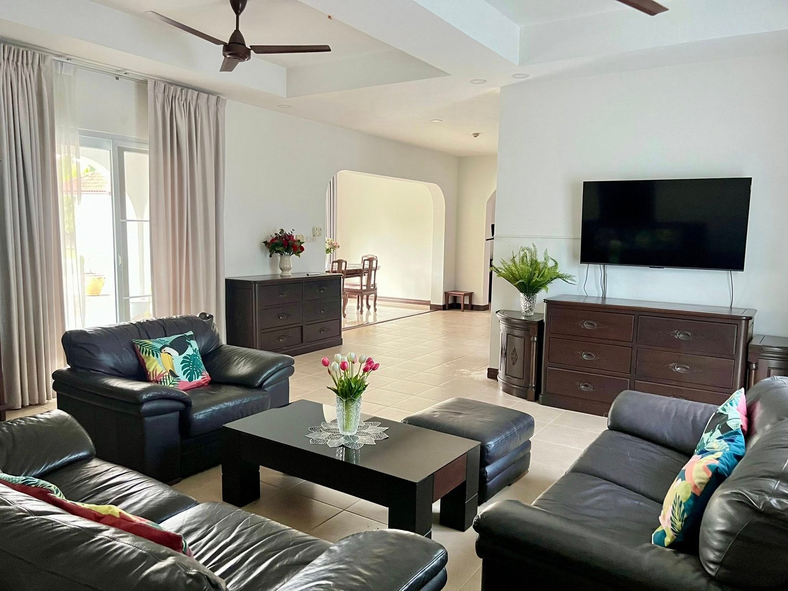 House_Sale_Mabprachan_Pattaya_Dlh533_3 Bright living room with black leather sectional, glass-top coffee table, and multiple throw pillows; TV on wall to the right, plants and dark wood furniture throughout.