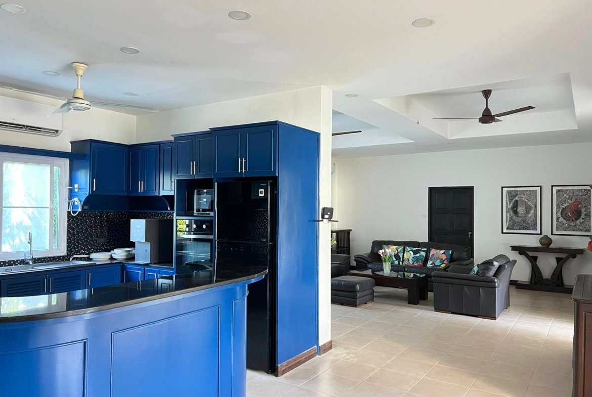 Open-concept kitchen with blue cabinets and island, stainless appliances, and a adjacent living area with dark seating and wall art in the background.