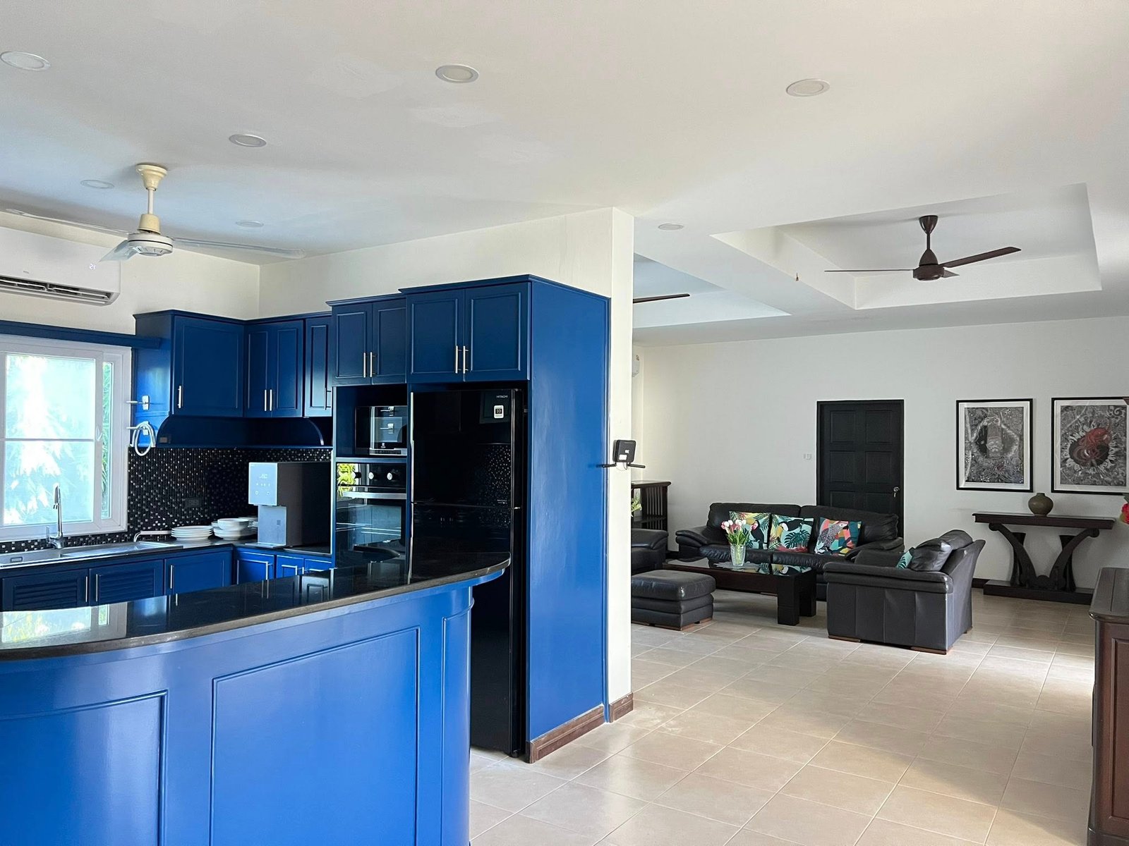 House_Sale_Mabprachan_Pattaya_Dlh533_4 Open-concept kitchen with blue cabinets and island, stainless appliances, and a adjacent living area with dark seating and wall art in the background.