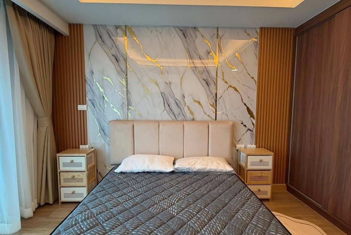 Hotel-style bedroom with an upholstered headboard, marble feature wall, and two wooden nightstands beside a double bed with a dark quilted coverlet.