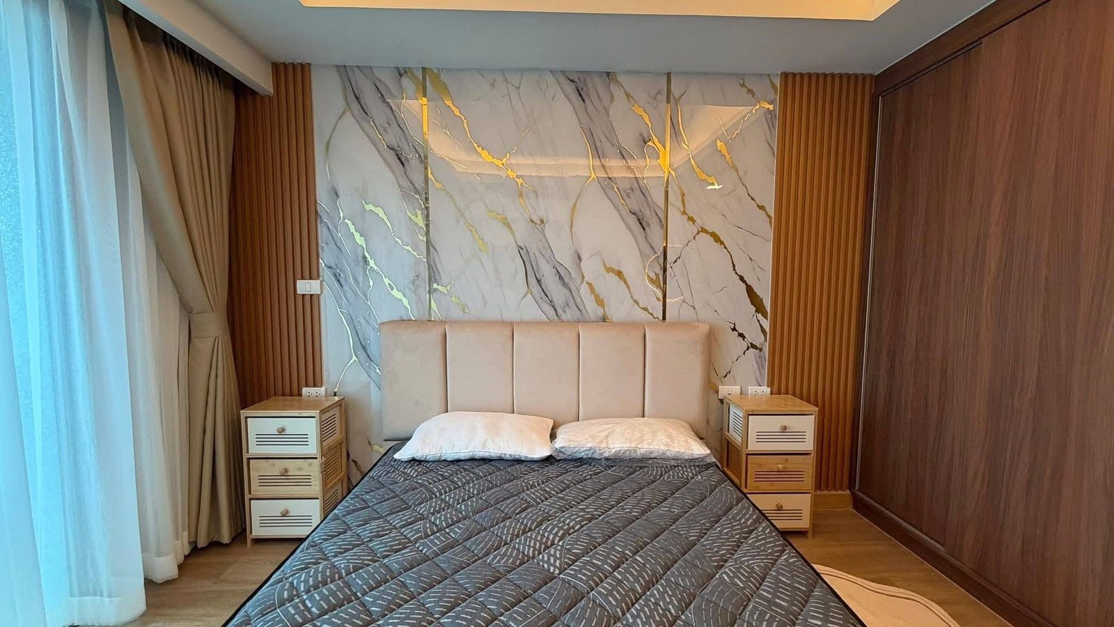 LBR2_Pattaya_Sale_Dlc613 Hotel-style bedroom with an upholstered headboard, marble feature wall, and two wooden nightstands beside a double bed with a dark quilted coverlet.