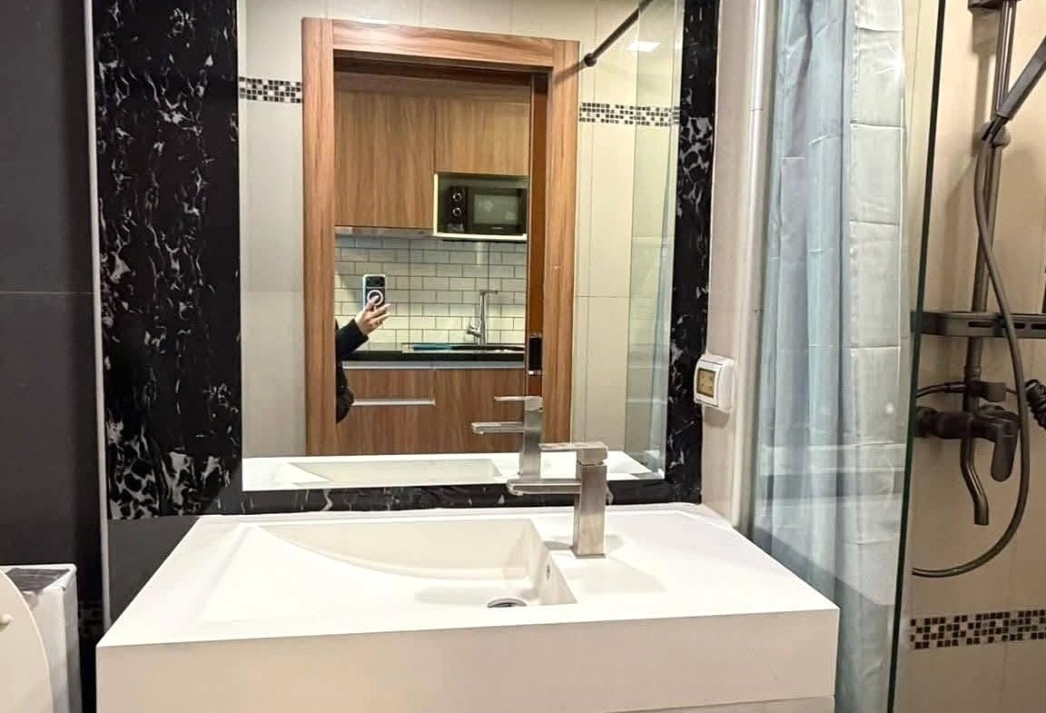 White rectangular bathroom vanity with an open lower shelf and integrated sink, mirrored by a large frame surrounded by black marble, with a glass shower enclosure to the right