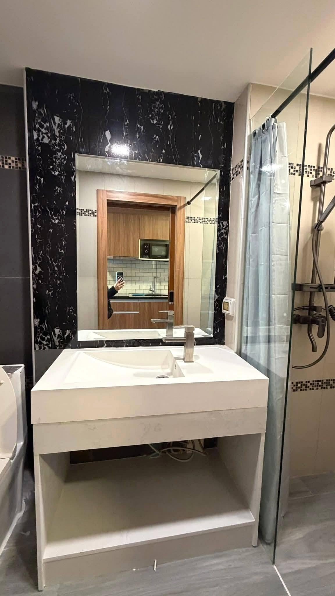 LBR2_Pattaya_Sale_Dlc613_10 White rectangular bathroom vanity with an open lower shelf and integrated sink, mirrored by a large frame surrounded by black marble, with a glass shower enclosure to the right