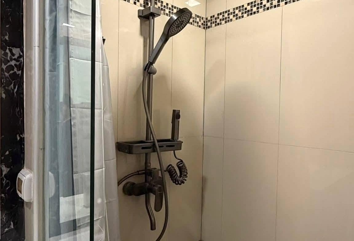 Modern shower stall with a large ceiling-mounted rainfall showerhead, a handheld shower on a chrome rail, glass partition, and white tiled walls with a black mosaic stripe intertile border.