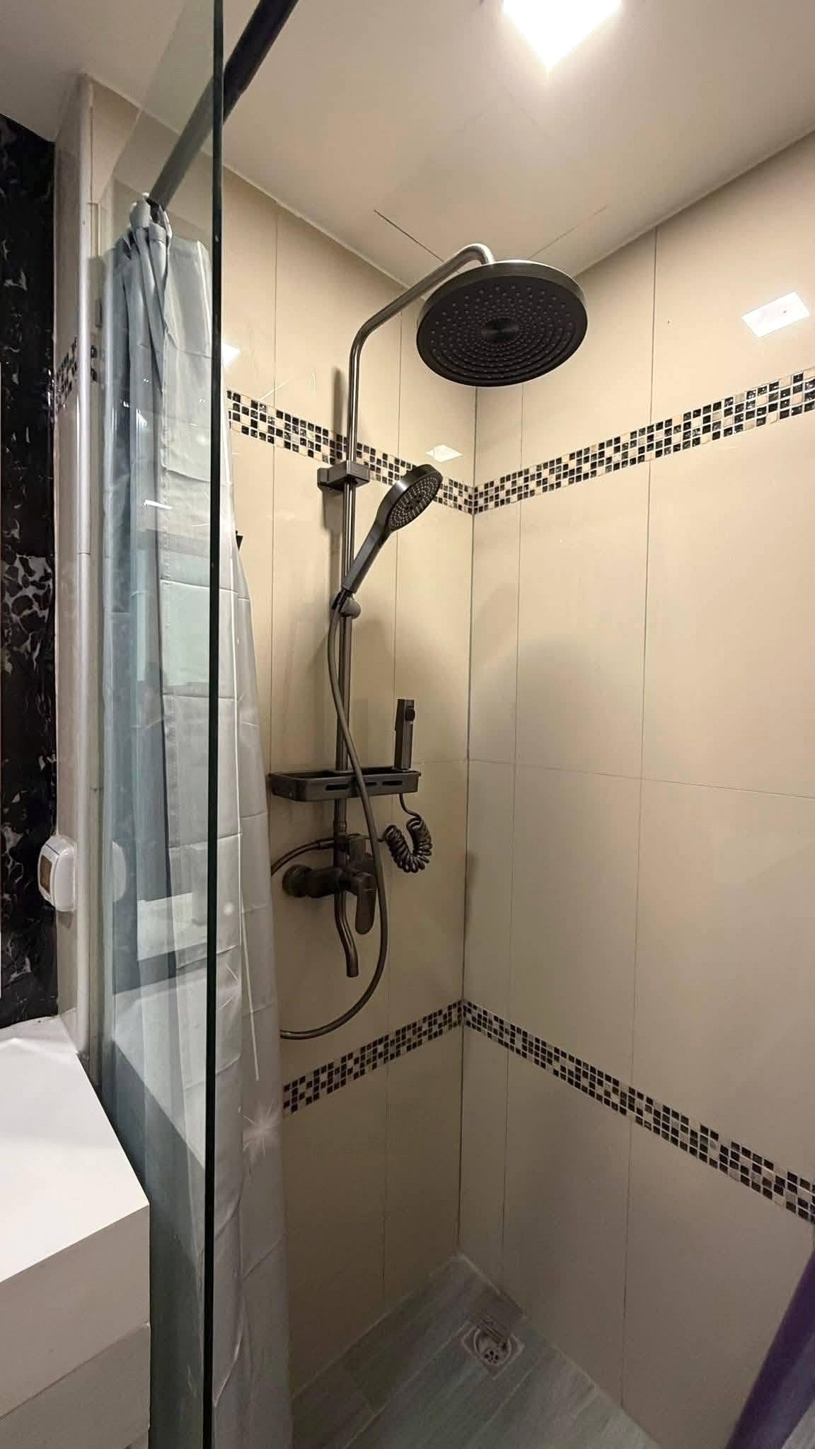 LBR2_Pattaya_Sale_Dlc613_11 Modern shower stall with a large ceiling-mounted rainfall showerhead, a handheld shower on a chrome rail, glass partition, and white tiled walls with a black mosaic stripe intertile border.