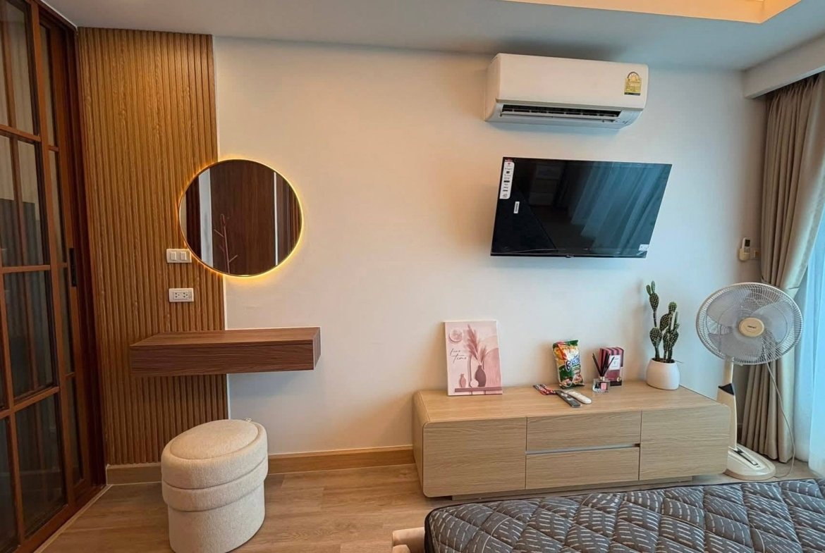 Modern hotel room corner with a round backlit mirror, wall-mounted TV, and light wood dresser beneath.