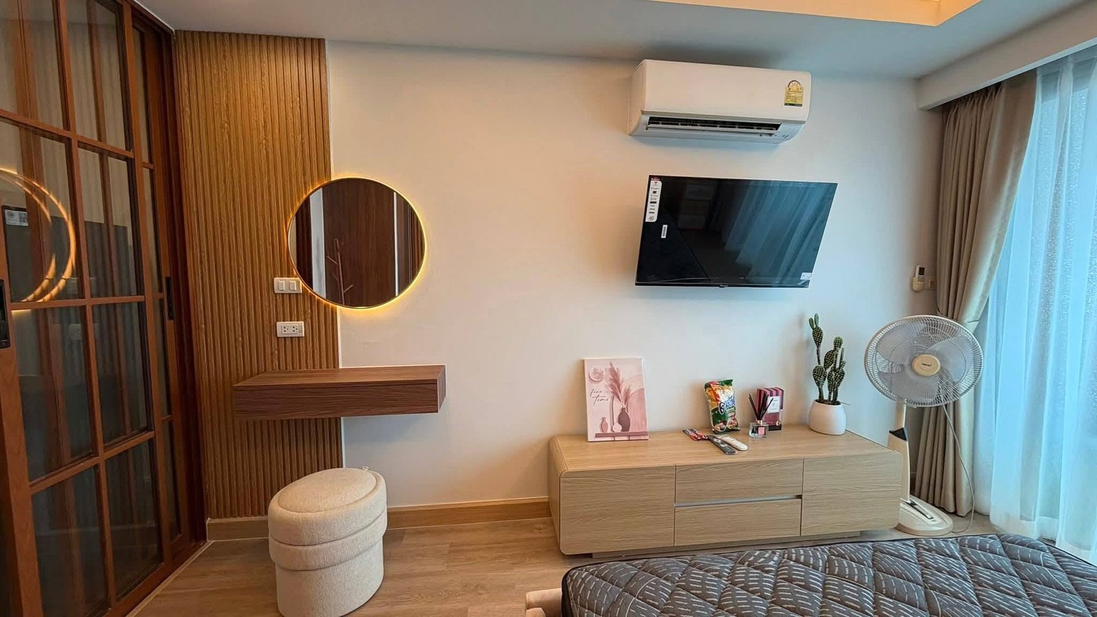 LBR2_Pattaya_Sale_Dlc613_2 Modern hotel room corner with a round backlit mirror, wall-mounted TV, and light wood dresser beneath.