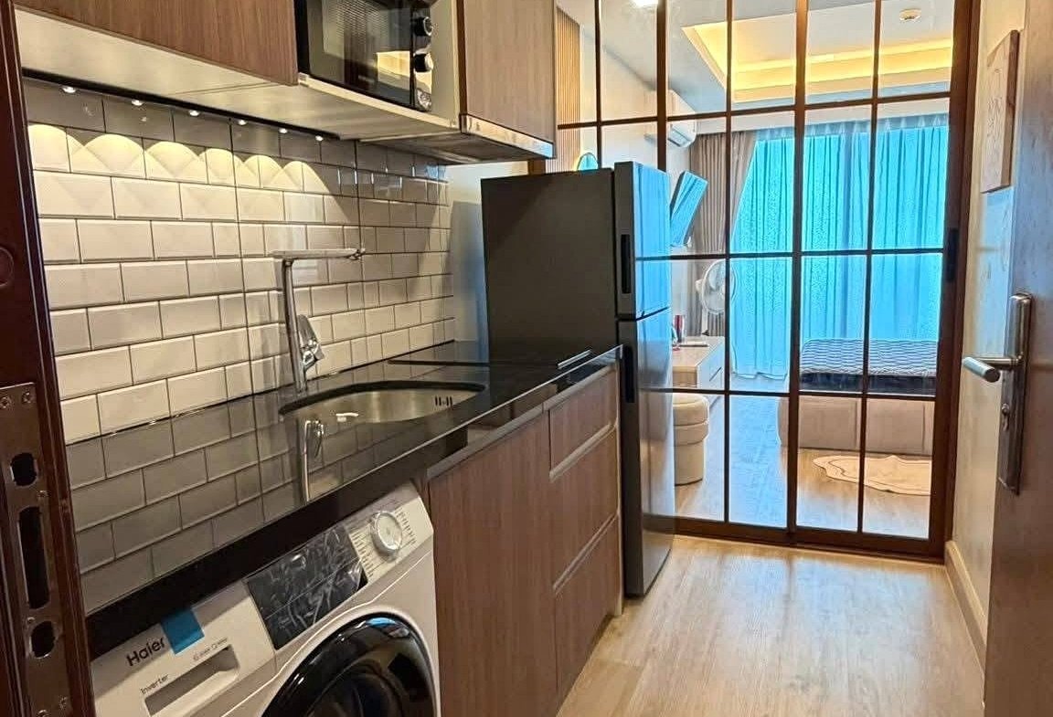 Modern galley kitchen with wooden cabinets, stainless appliances, sink, and a glass‑panel door opening to a living area with blue curtains.