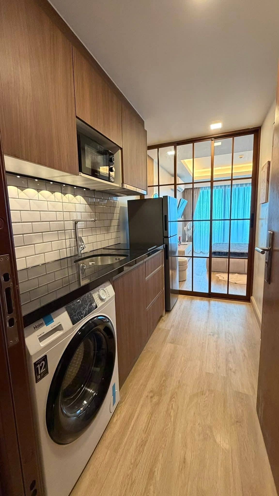 LBR2_Pattaya_Sale_Dlc613_3 Modern galley kitchen with wooden cabinets, stainless appliances, sink, and a glass‑panel door opening to a living area with blue curtains.