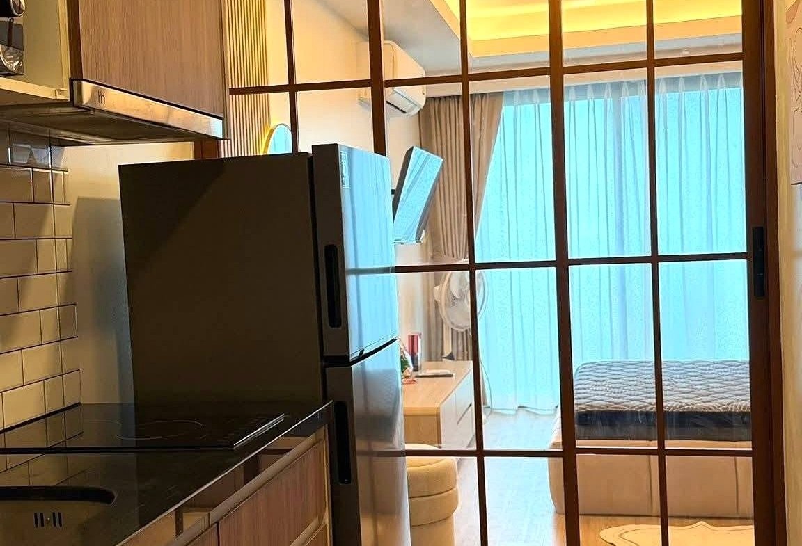 Modern kitchen with wooden cabinets and a stainless steel refrigerator, opening to a glass-panel partition leading to a bright living area.