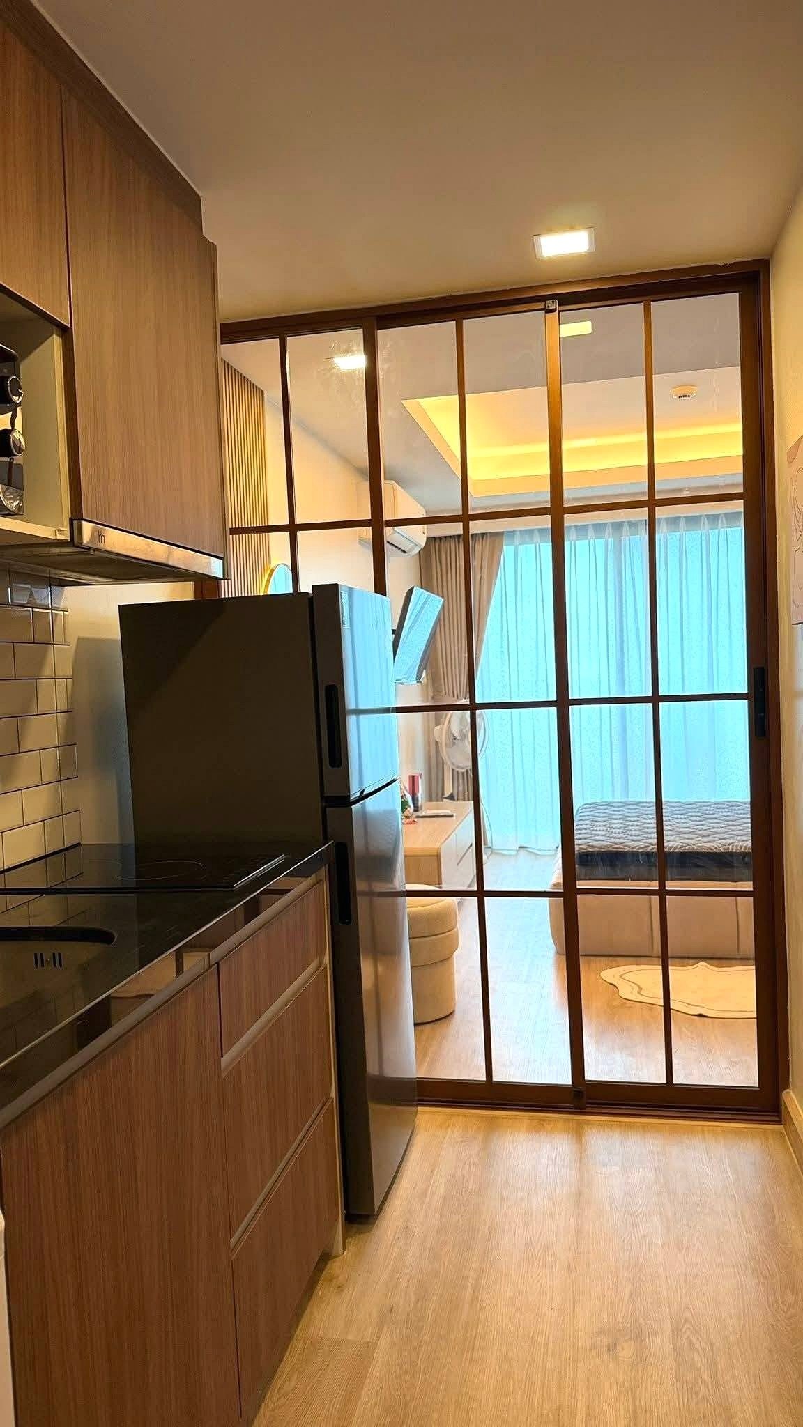 LBR2_Pattaya_Sale_Dlc613_4 Modern kitchen with wooden cabinets and a stainless steel refrigerator, opening to a glass-panel partition leading to a bright living area.