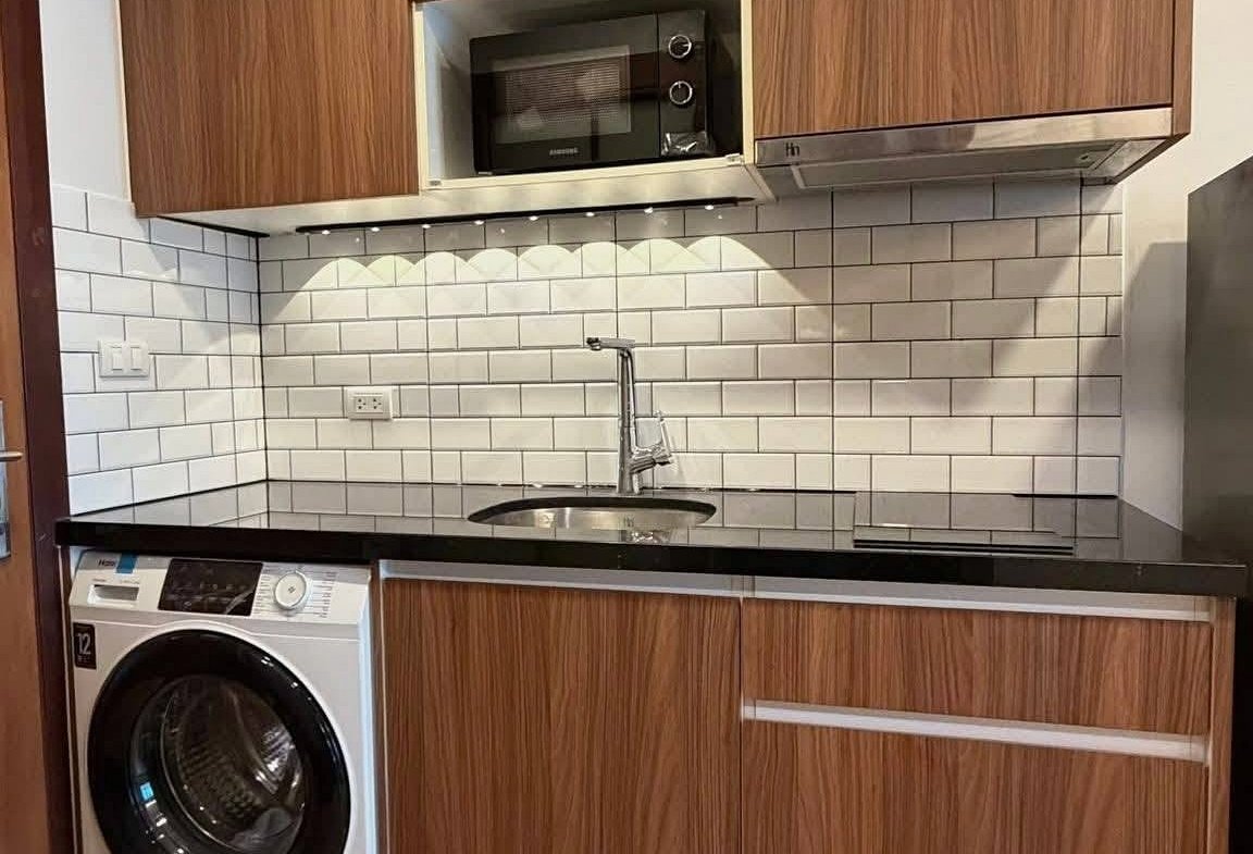 Modern kitchen with wooden cabinets, stainless steel countertop, microwave, white subway tile backsplash, and a front-loading washing machine on the left.